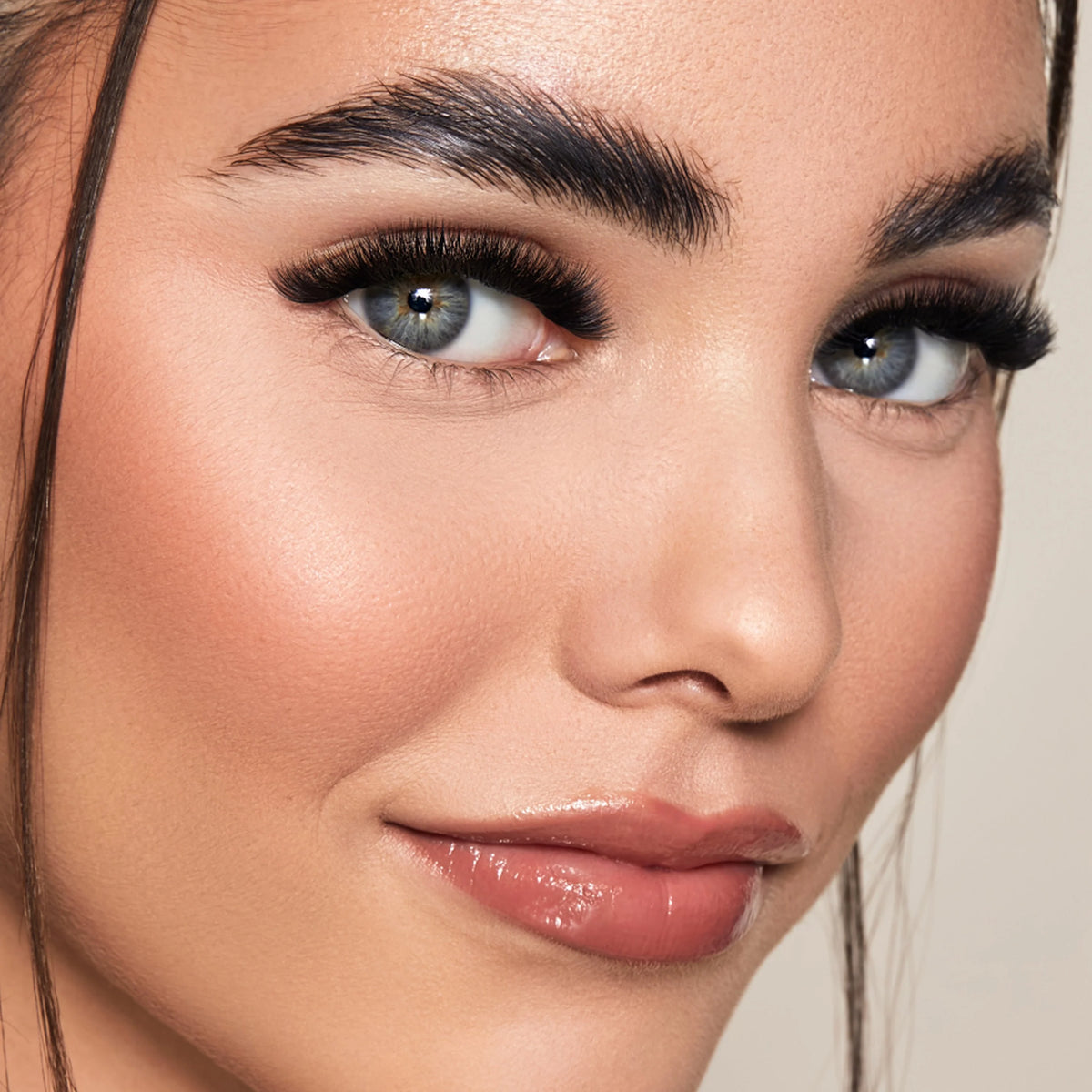 Close-up of a woman's face showcasing long, dark, curled false eyelashes and a glossy nude-pink lip gloss against a soft beige background. Showcasing what can be created with Lash Box LA Australia Education and supplies designed to save you time